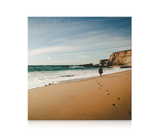 Ein quadratischer Leinwanddruck einer Frau, die am Strand spazieren geht, hängt an einer Wand.