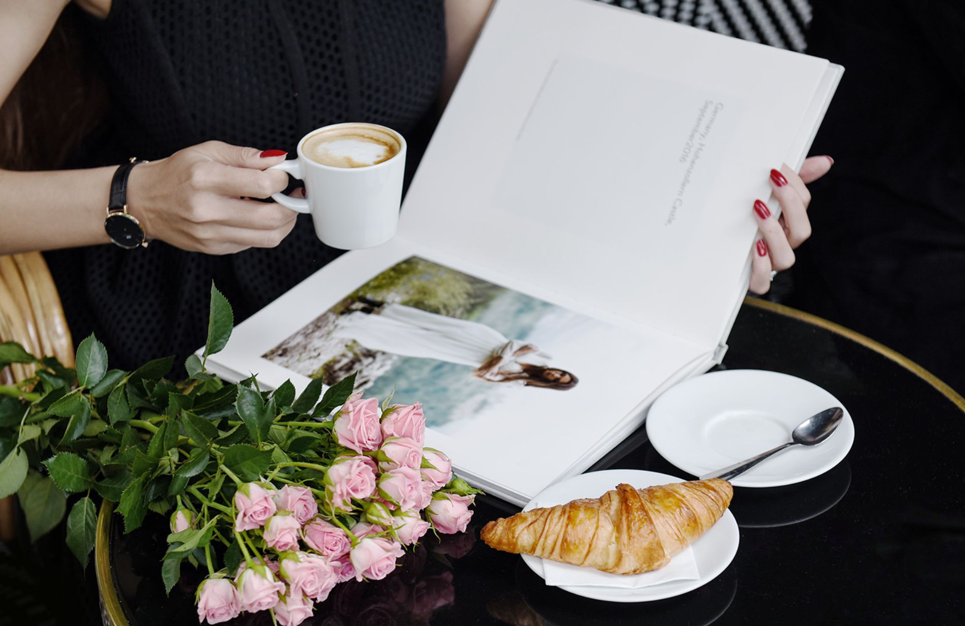 Woman looking through a portrait photo book at a table with coffee, roses and croissant.
