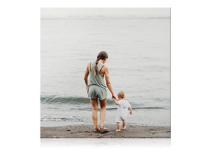 Square canvas print of mother and her child holding hands and dipping their feet in the water at the beach.