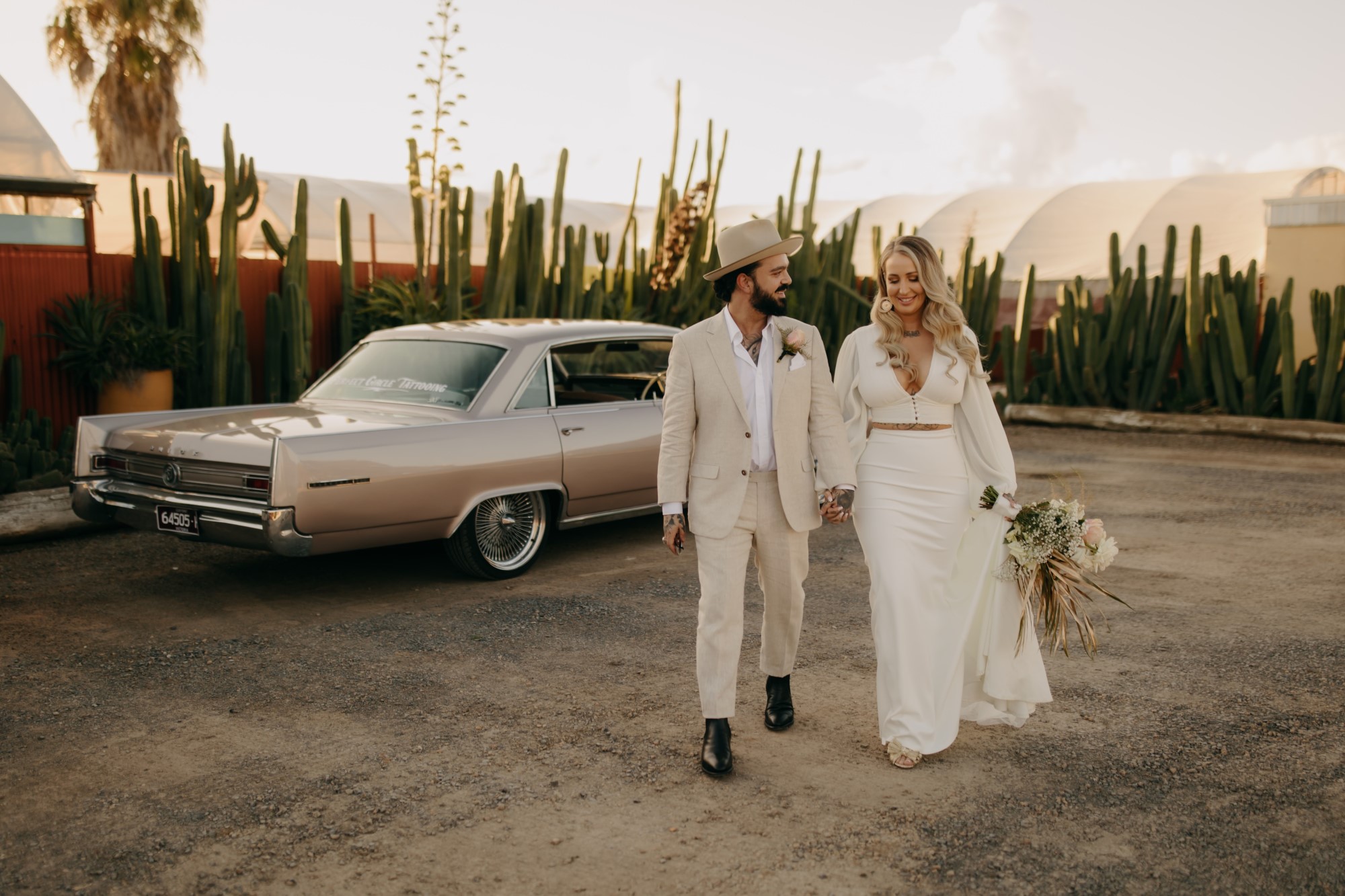 Bridal couple holding hands and walking in front of a vintage car in the desert with surrounding cacti