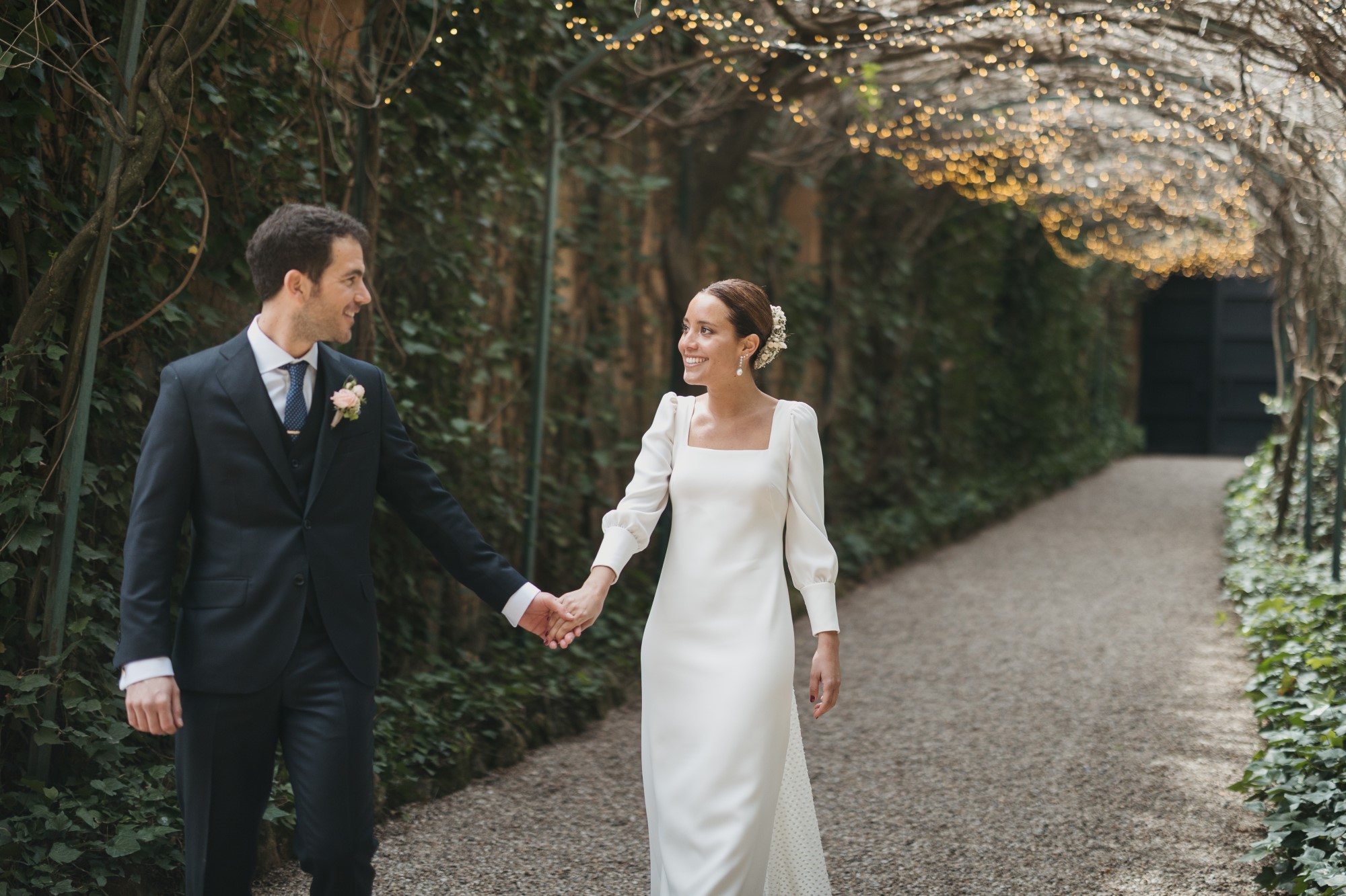 Bridal couple at wedding holding hands and walking together under floral archway with string lights