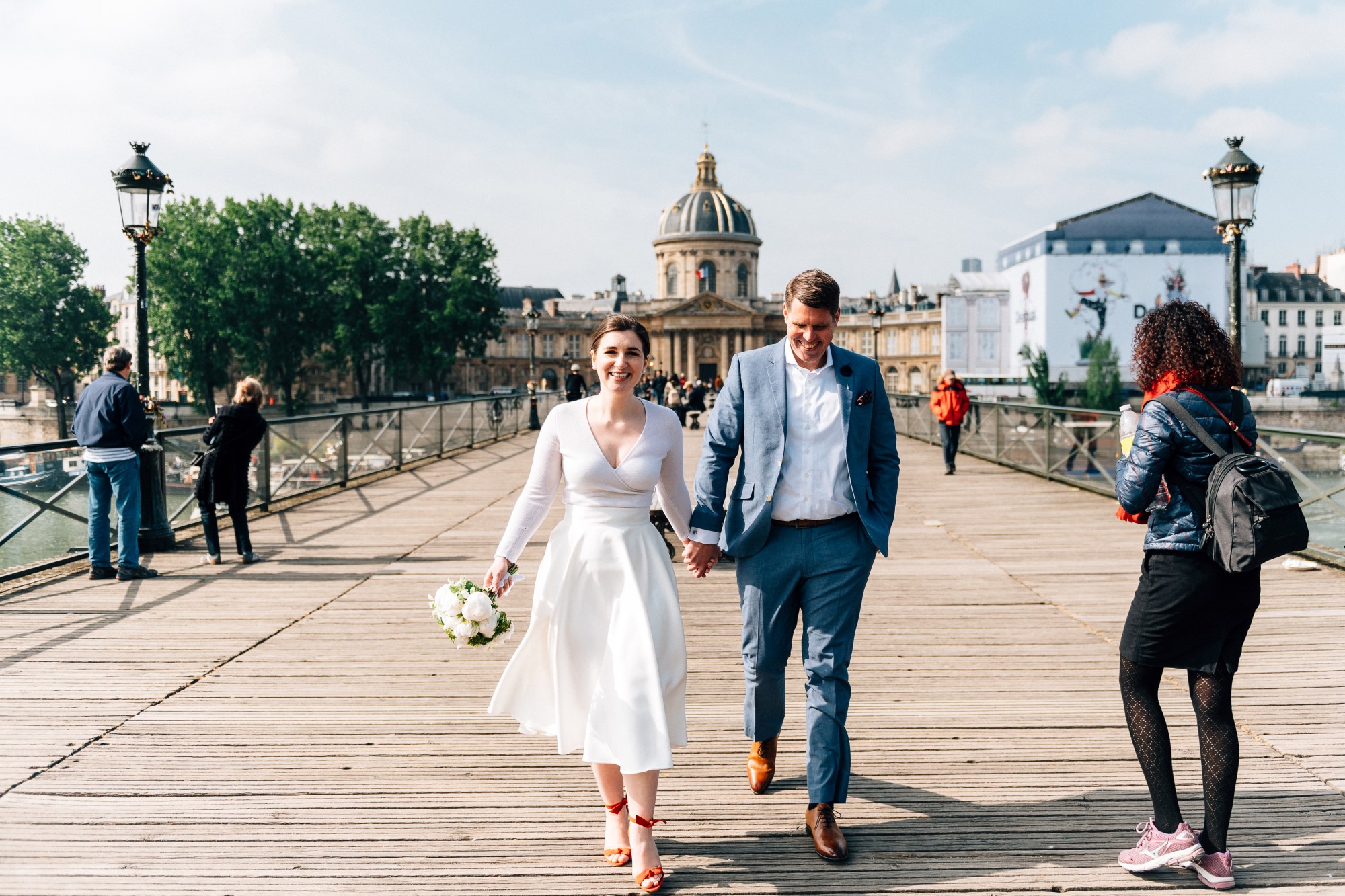 Bridal couple holding hands together in Paris on a bridge, bride wears vintage white tea-length dress with red shoes