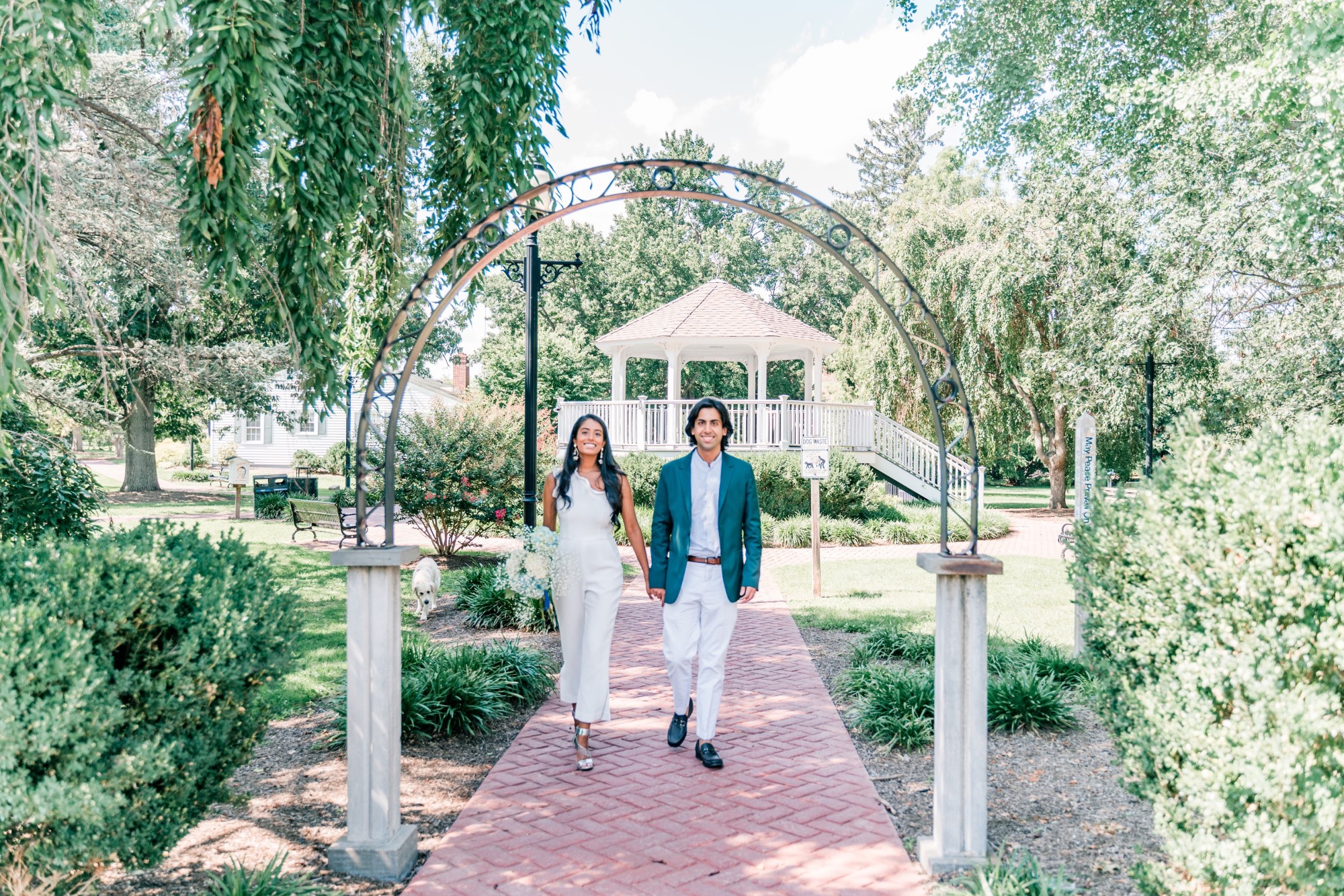 Bridal couple facing and walking towards camera, bride wears white pant suit and holds a flower bouquet