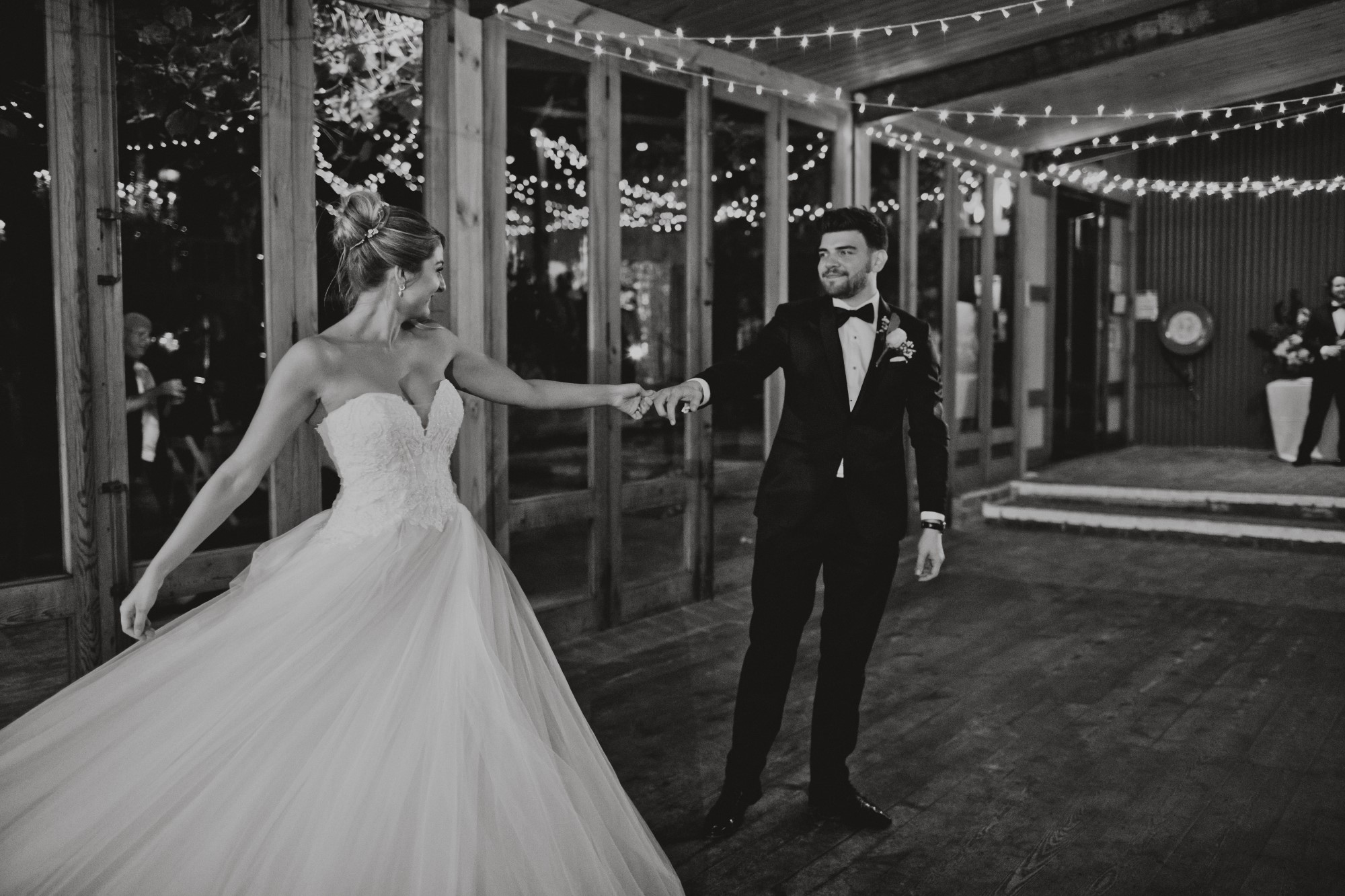 Black and white photo of bride and groom dancing together at a wedding reception with string lights above them