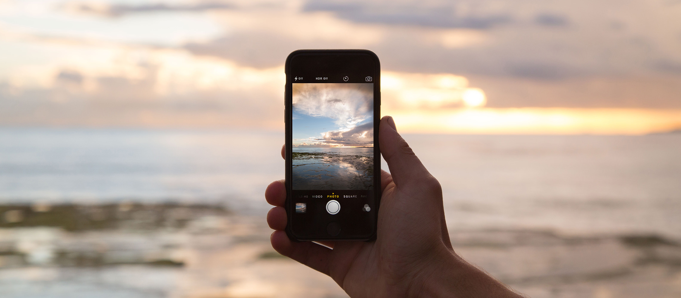 Hand holding iPhone camera taking a photo of a sunset at the beach.