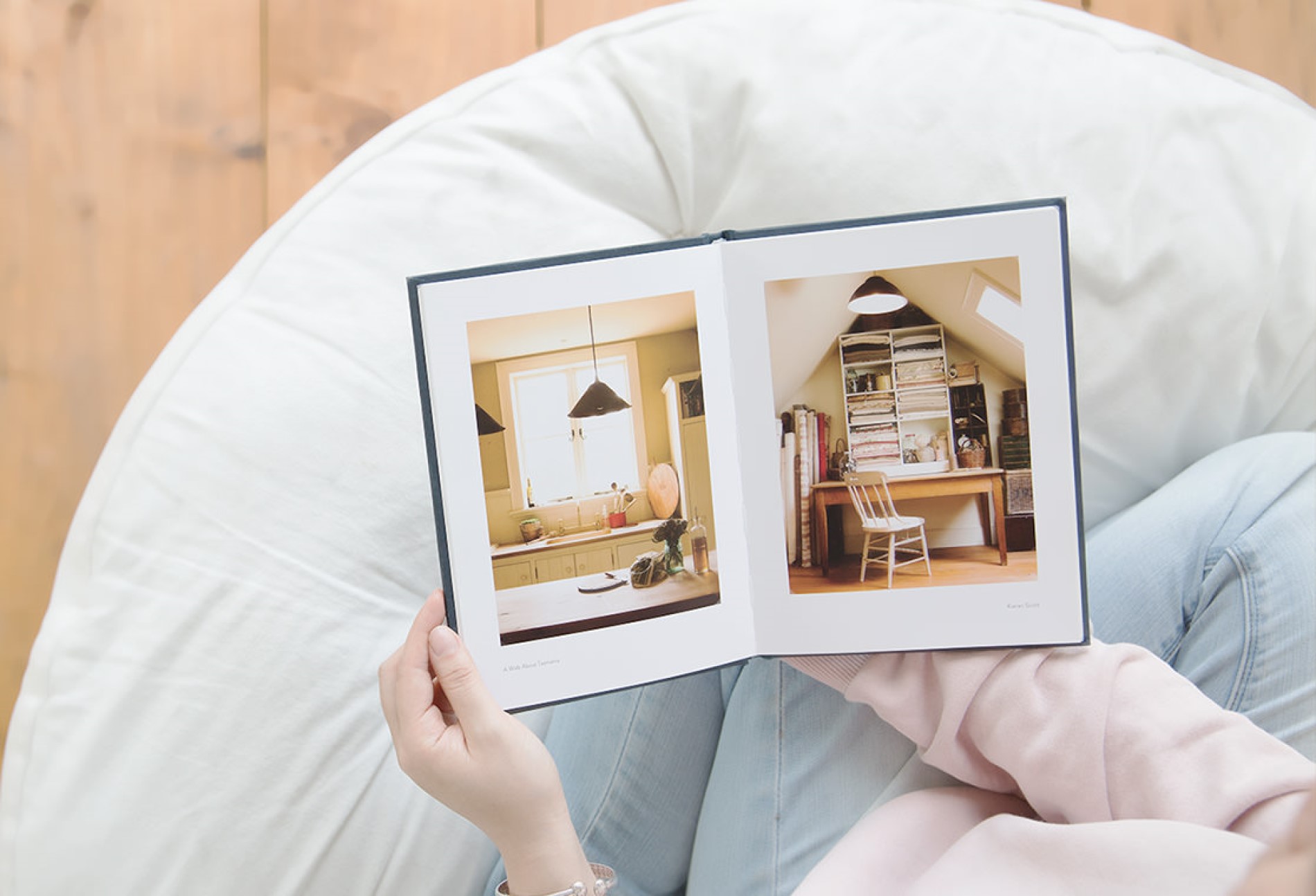 Over the shoulder shot of a female in a pink jumper and blue jeans holding open a photo book containing images of home interiors.