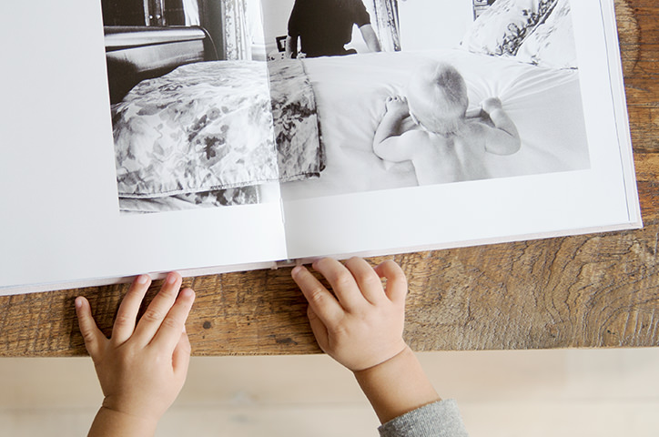 Ein Kleinkind blättert durch ein quadratisches Fotobuch mit Bildern von lachenden jungen Mädchen.