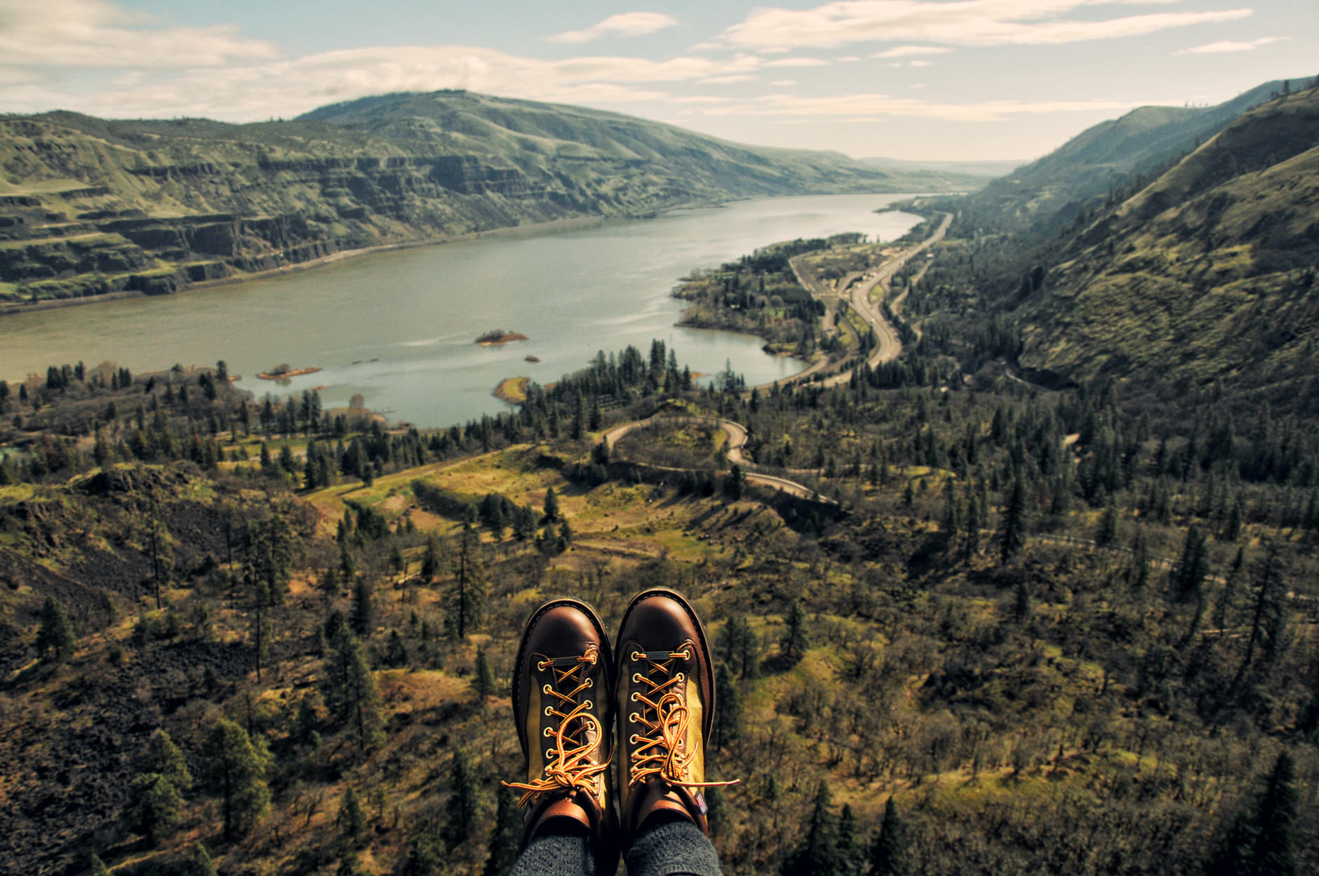 A person's boots dangling with a forest and sea view in the background.