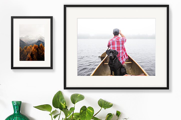 Two hanging gallery frames side by side on a white wall; one is an image of autumnal trees, the other is a man and his dog on a canoe.