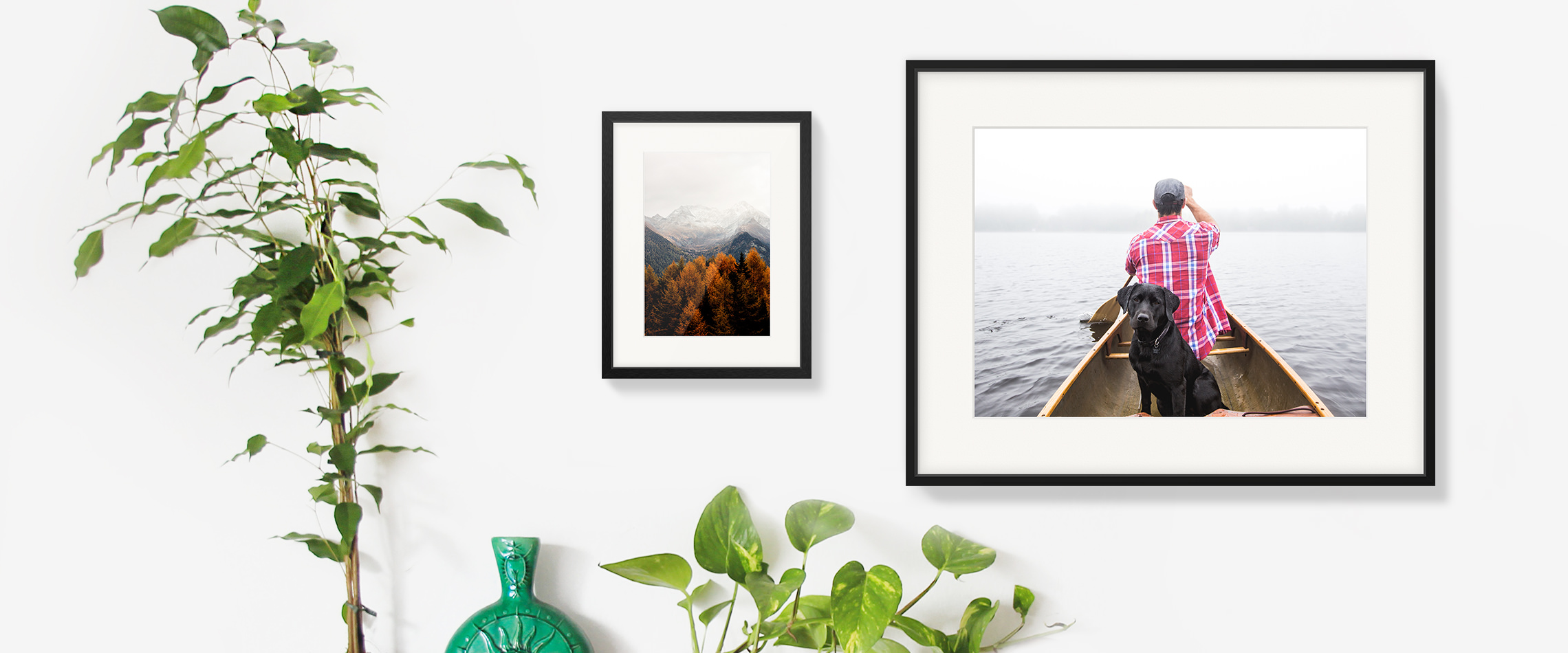 Two hanging gallery frames side by side on a white wall; one is an image of autumnal trees, the other is a man and his dog on a canoe.