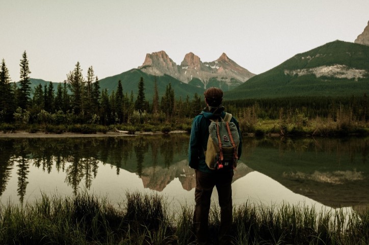 Mann steht am Seeufer und schaut auf Berge und Wald in der Ferne.