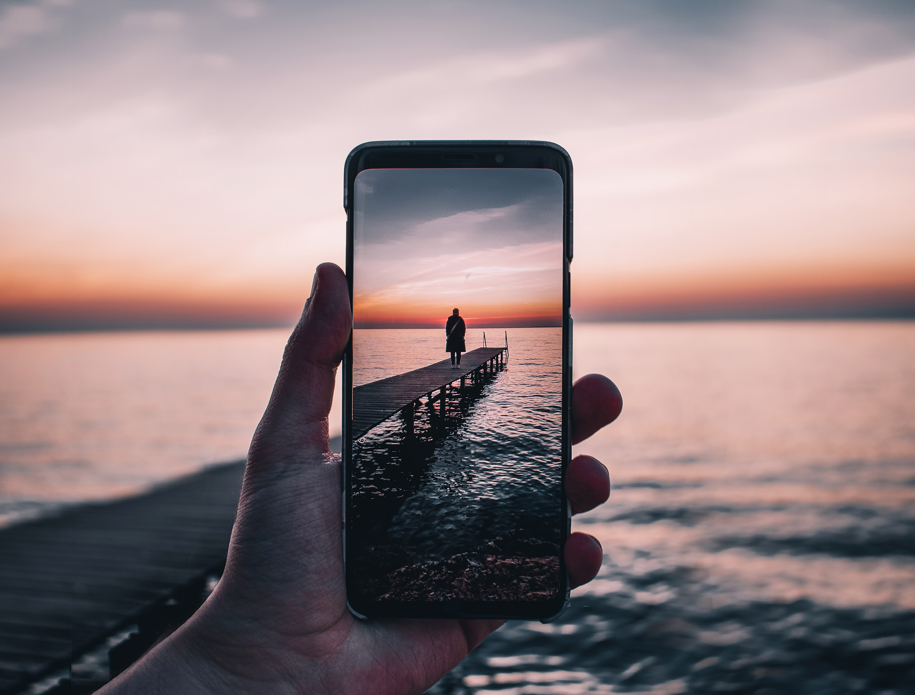 Mano che tiene iPhone nella vista della fotocamera con una persona sul molo al tramonto.