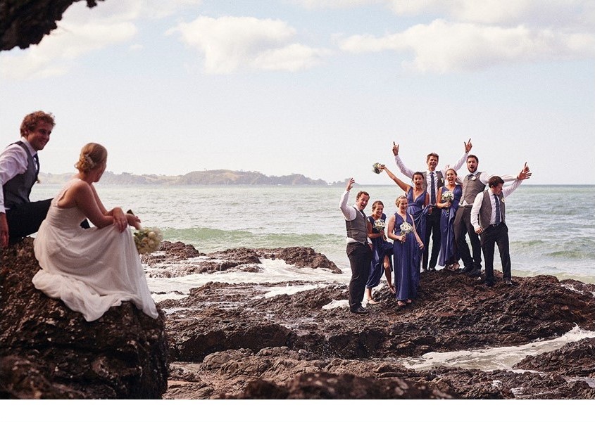 Bride and groom watch as wedding party pose for photo by sea