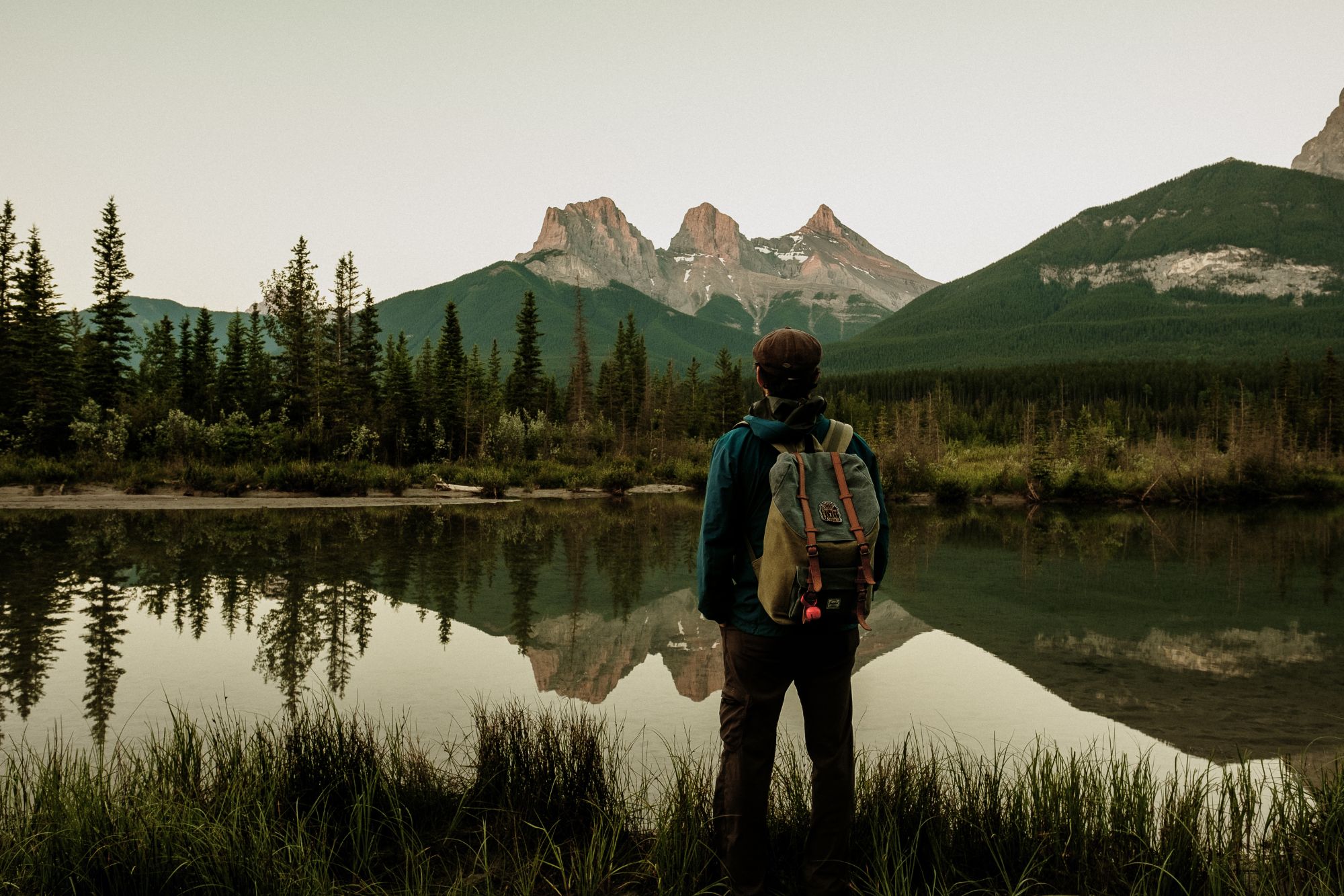Male standing lakeside looking at mountains and forest in distance.