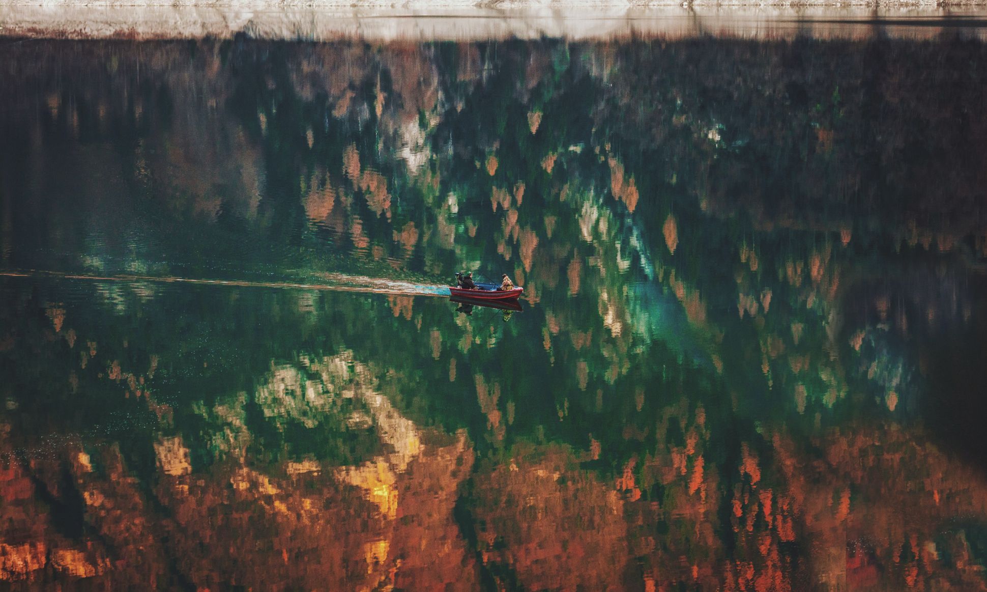Two people in a small boat moving across water with reflection of autumn trees.