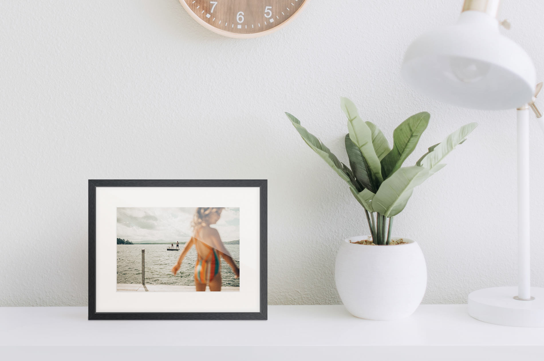 A gallery frame set on a white table, alongside a pot plant and white lamp.