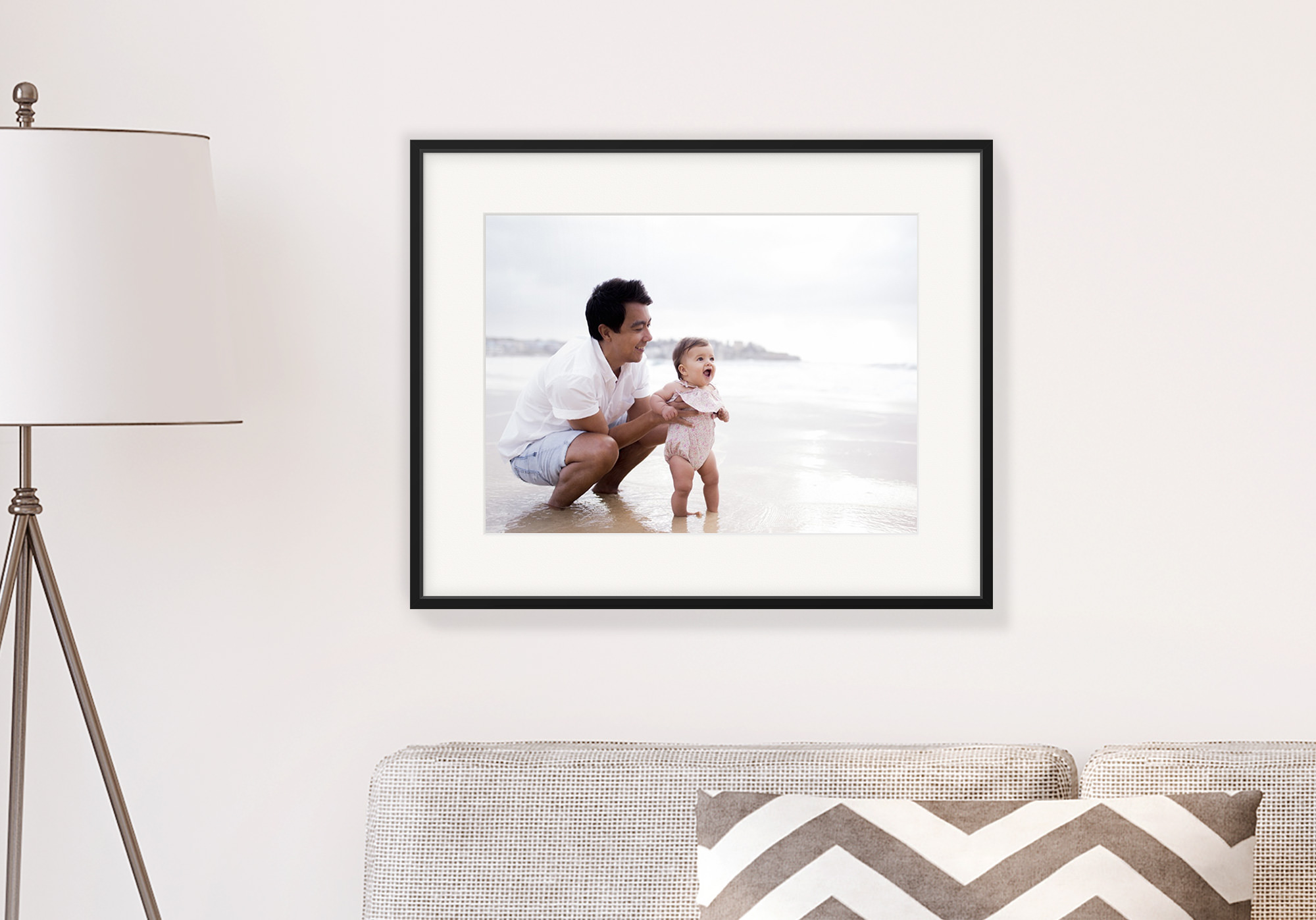 A photo frame hung on a wall behind a sofa and to the right of a standing lamp, with an image of a father and his young daughter paddling in the sea.