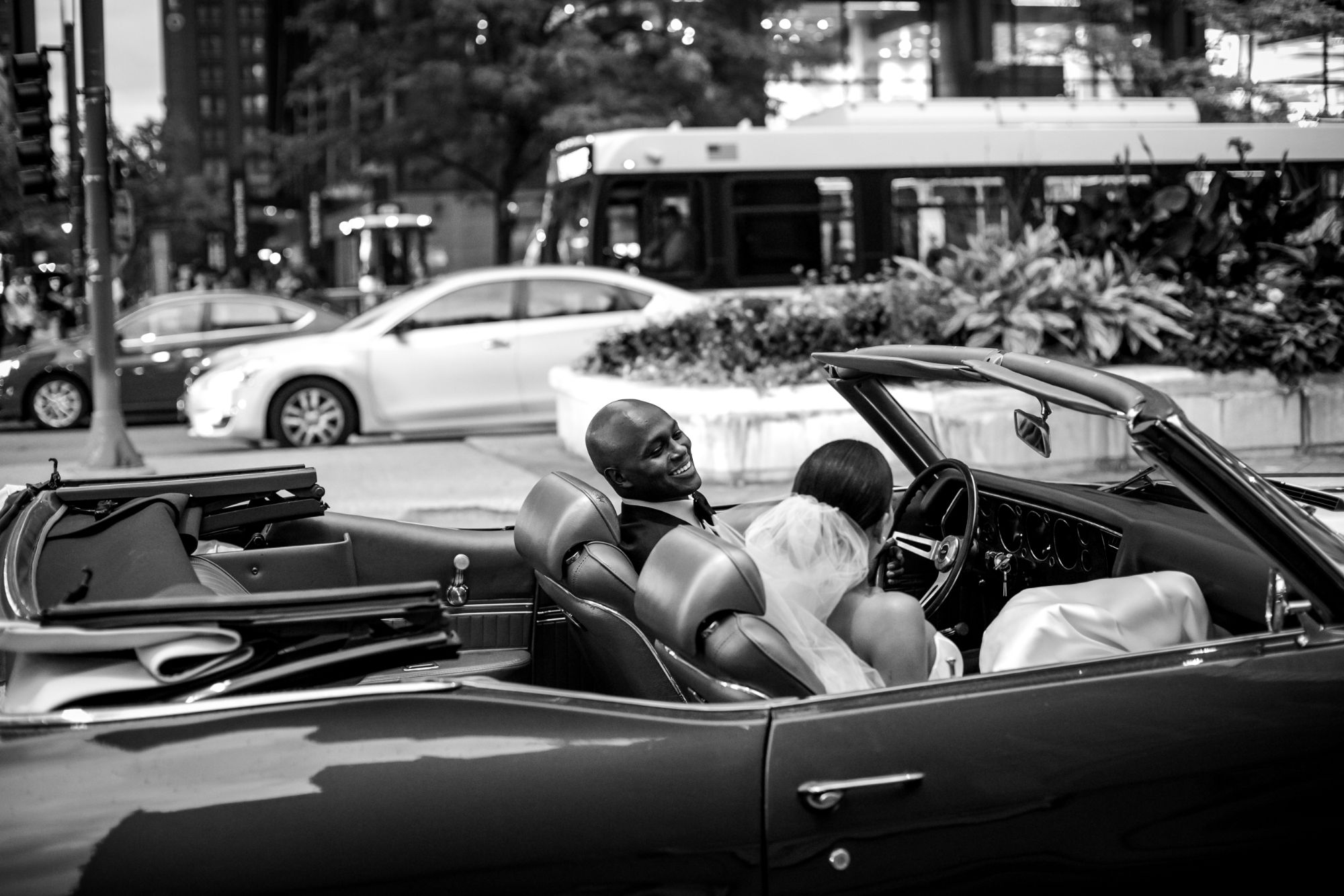 Groom smiling at his new bride in top-down convertible car.