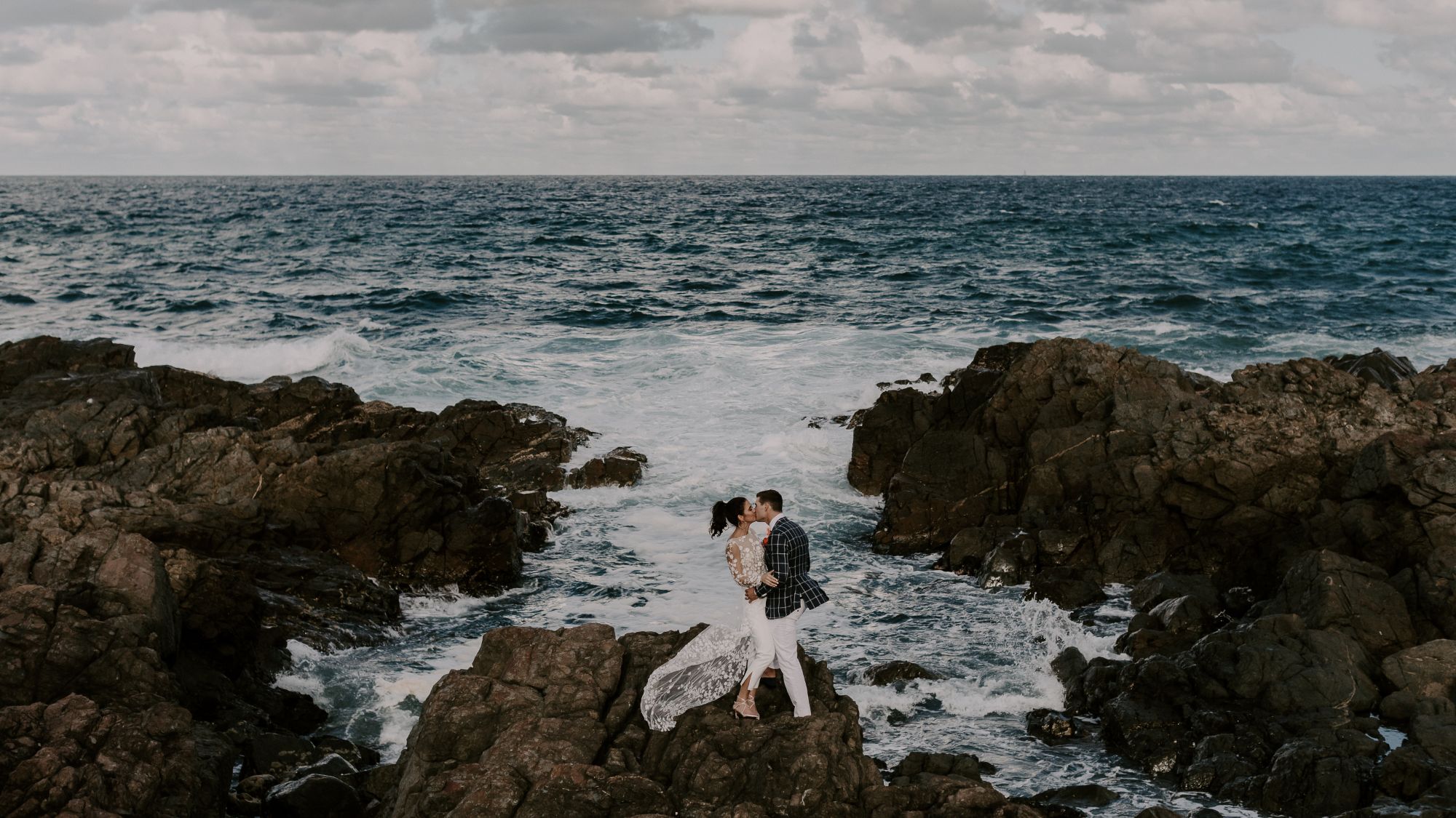 Newlywed couple kissing on the rocks next to sea.