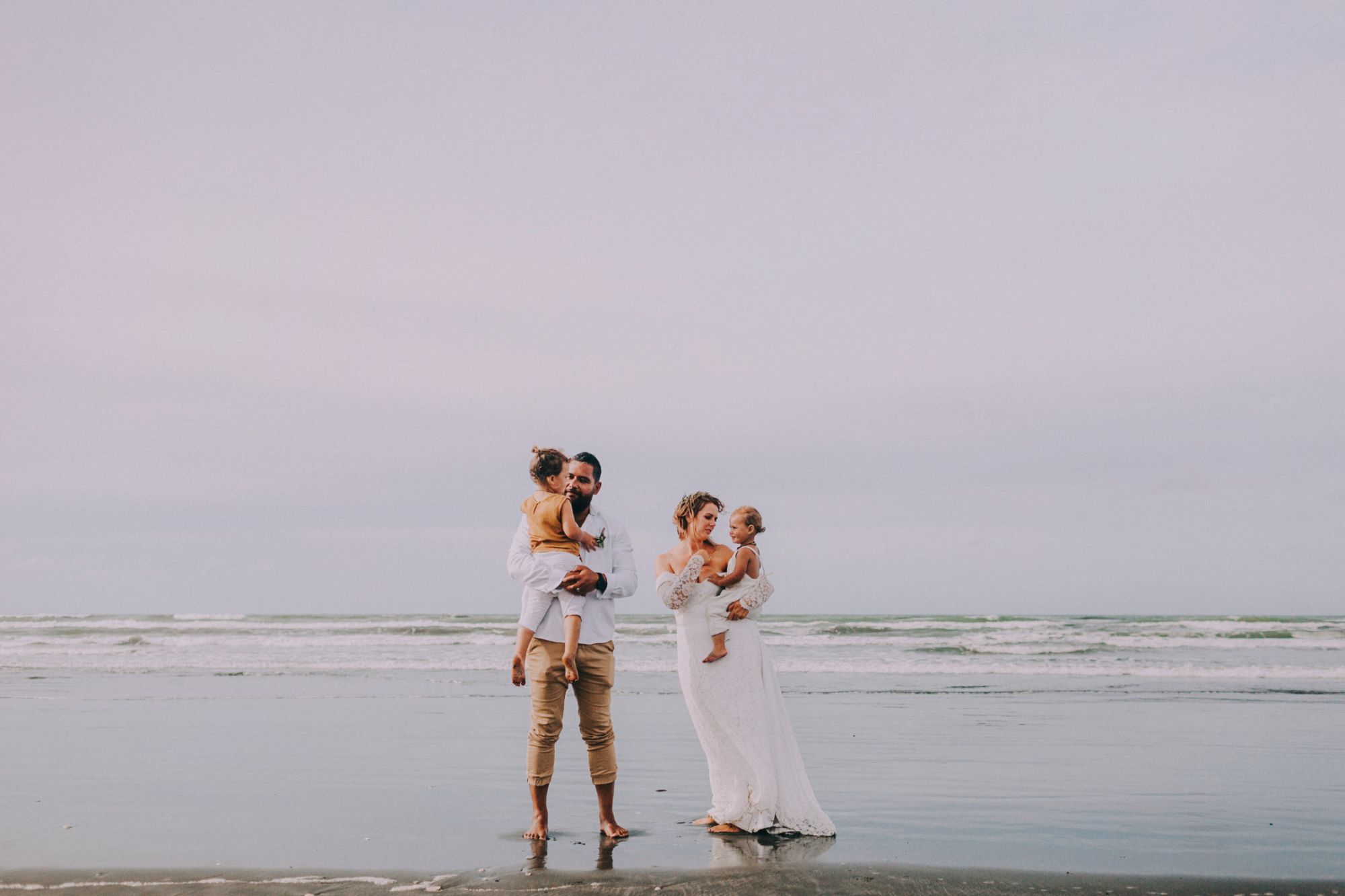 Newlywed couple holding two young children at the beach.