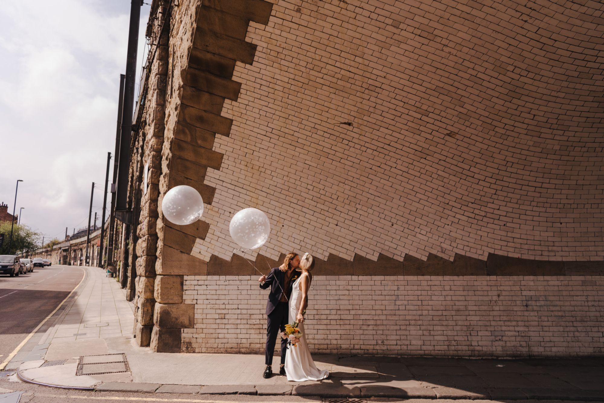 Newlywed couple holding balloons, kissing in brick tunnel.