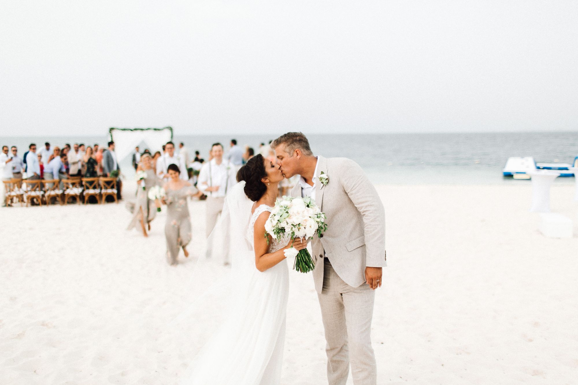 Newlywed couple kissing after beach ceremony with guests in background.
