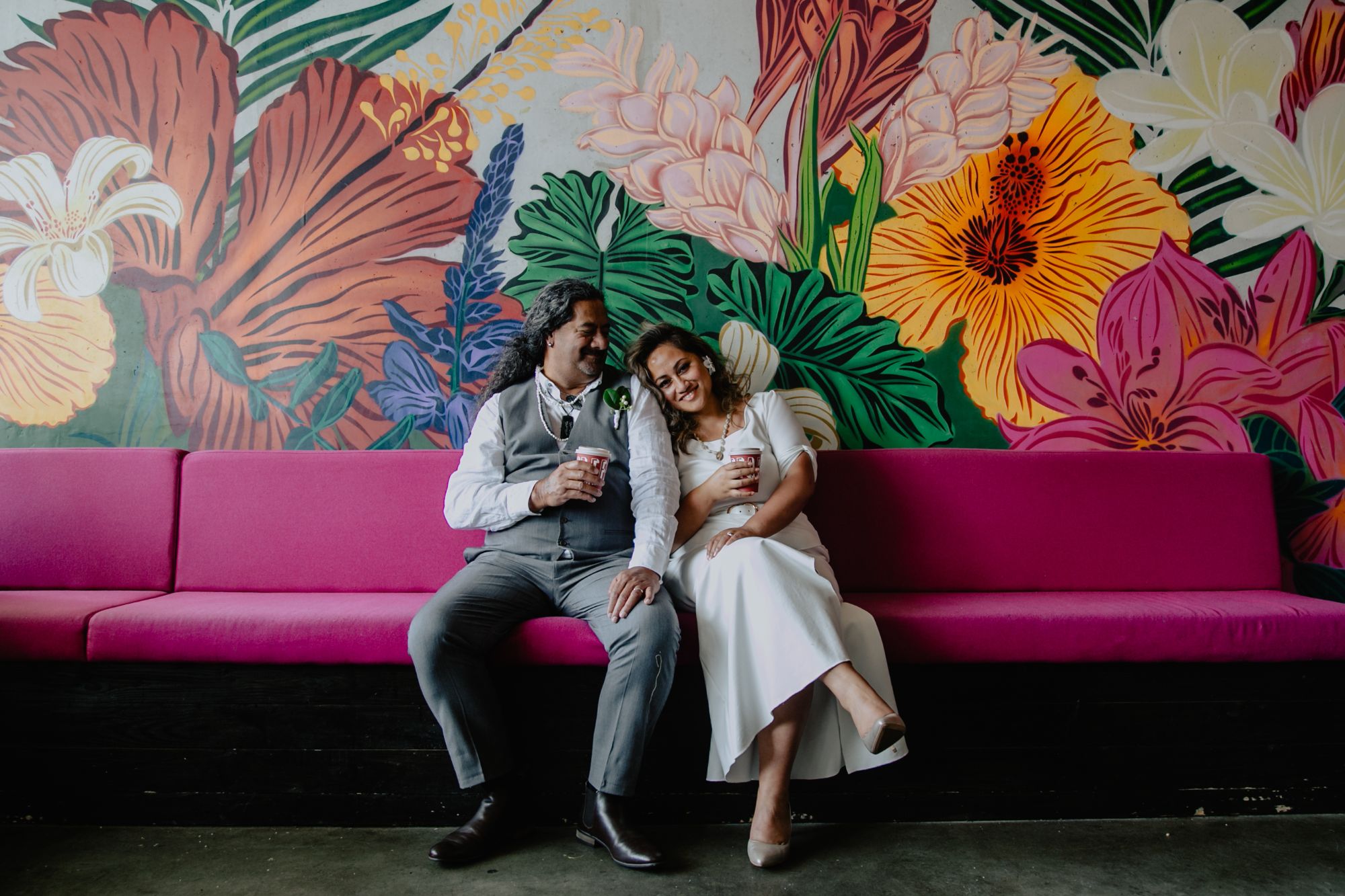 Newlywed bride and groom sitting on pink sofa with colorful floral painting on wall behind.