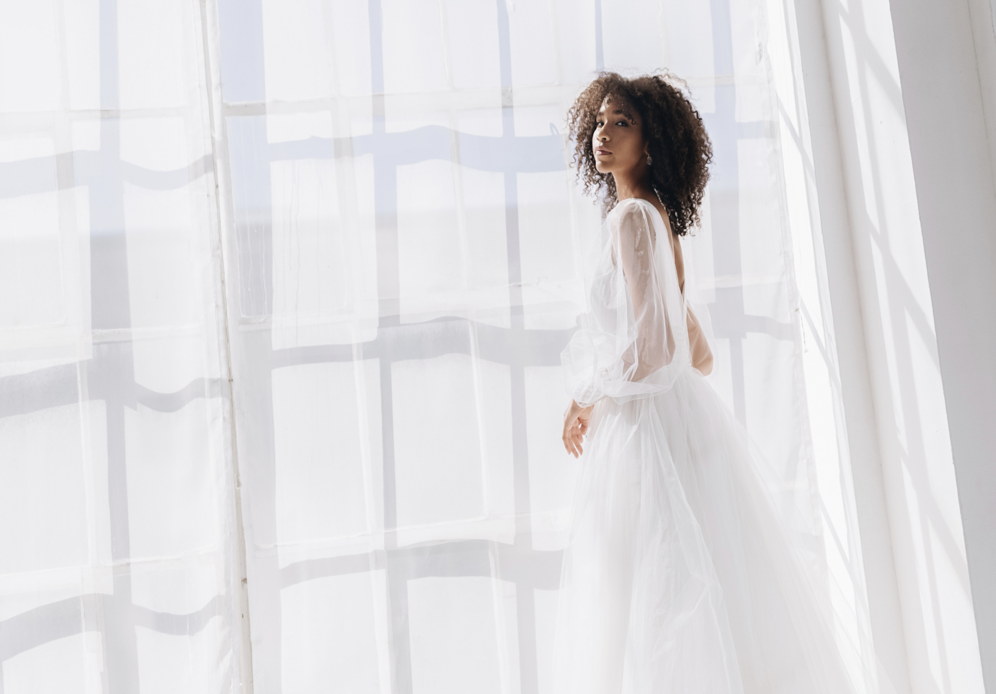 Mujer con vestido de novia blanco junto a una ventana.