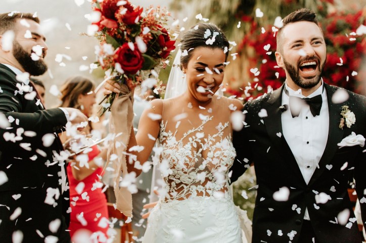 Happy newlyweds walking through white confetti as guests clap